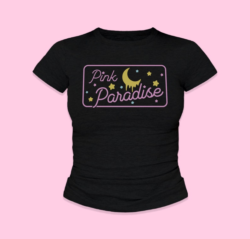 May include: A black t-shirt with a pink rectangle that says 'Pink Paradise' in a retro font. The rectangle has a yellow crescent moon with a dripping yellow liquid and yellow stars around it.