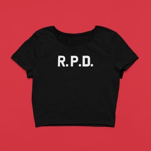 May include: Black crop top with the white letters "R.P.D." printed across the front. The shirt has short sleeves and a scoop neckline. The background is a solid red.