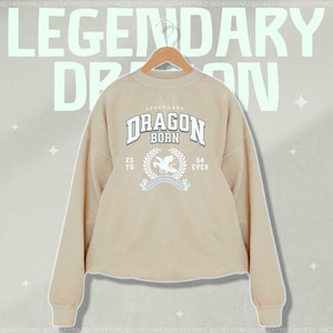 May include: A beige sweatshirt with a white graphic print of a dragon with the text "Legendary Dragon Born" and "Est. 04 Ever".