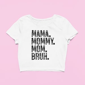 May include: A white short-sleeved crop top featuring black distressed-style text printed on the front. The text reads MAMA. MOMMY. MOM. BRUH. in a stacked, sans-serif font. The garment has a wide, rounded neckline and a cropped hemline.