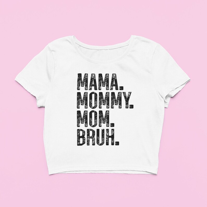 May include: A white short-sleeved crop top featuring black distressed-style text printed on the front. The text reads MAMA. MOMMY. MOM. BRUH. in a stacked, sans-serif font. The garment has a wide, rounded neckline and a cropped hemline.