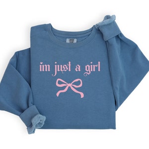 Op de afbeelding: Een blauw sweatshirt met de tekst "im just a girl" in roze gotische letters en een roze strik eronder.