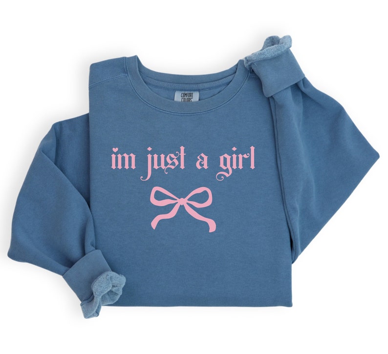 Op de afbeelding: Een blauw sweatshirt met de tekst "im just a girl" in roze gotische letters en een roze strik eronder.