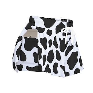 May include: A white athletic skort featuring a black cow print pattern. The garment has an elastic waistband with a white drawstring and a side pocket containing a light-colored smartphone. The design includes a flared skirt overlay.