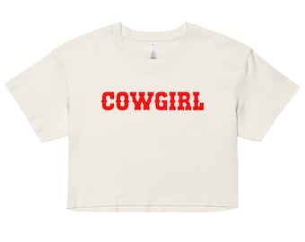 Puede incluir: Top corto color crema con la palabra "COWGIRL" impresa en letras rojas en negrita. La camiseta tiene mangas cortas y cuello redondo. El texto tiene una fuente de estilo occidental.