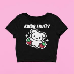 Op de afbeelding: Zwarte crop top met de tekst "Kinda Fruity" boven een wit cartoonfiguur dat twee rode aardbeien vasthoudt. Het ontwerp bevat witte sterren. De top heeft korte mouwen en een ronde hals.