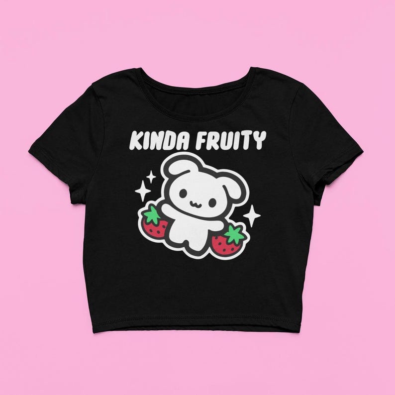 Op de afbeelding: Zwarte crop top met de tekst "Kinda Fruity" boven een wit cartoonfiguur dat twee rode aardbeien vasthoudt. Het ontwerp bevat witte sterren. De top heeft korte mouwen en een ronde hals.
