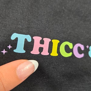 Op de afbeelding: Zwart T-shirt met het woord "THICC" in kleurrijke, bubbelletters. De letters zijn lichtblauw, roze, geel, groen en paars. Er staan kleine paarse sterren links van de tekst.