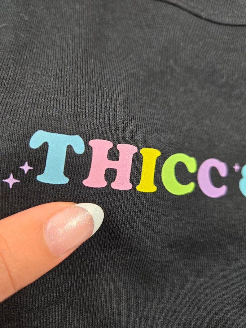 Op de afbeelding: Zwart T-shirt met het woord "THICC" in kleurrijke, bubbelletters. De letters zijn lichtblauw, roze, geel, groen en paars. Er staan kleine paarse sterren links van de tekst.