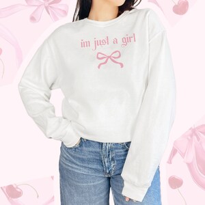 Op de afbeelding: Witte trui met de tekst "im just a girl" in roze letters en een roze strik.