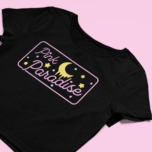 Pu&ograve; includere: T-shirt nero con una grafica rettangolare che recita "Pink Paradise" in corsivo. La grafica include una luna crescente gialla, stelle e un effetto gocciolante. La maglietta &egrave; ambientata su uno sfondo rosa.