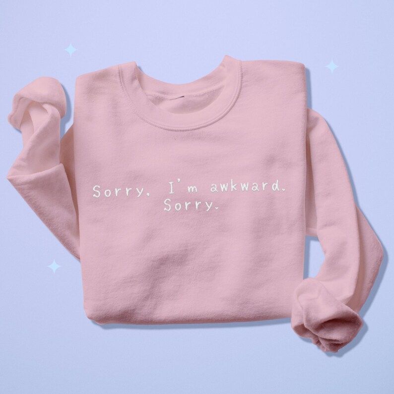 May include: A light pink sweatshirt with the text "Sorry. I'm awkward. Sorry." in white. The sweatshirt has long sleeves and a crew neck. The background is a light blue.