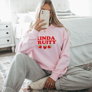 Puede incluir: Una sudadera rosa con el texto "Kinda Fruity" en letras rojas y tres fresas rojas impresas en la parte delantera.
