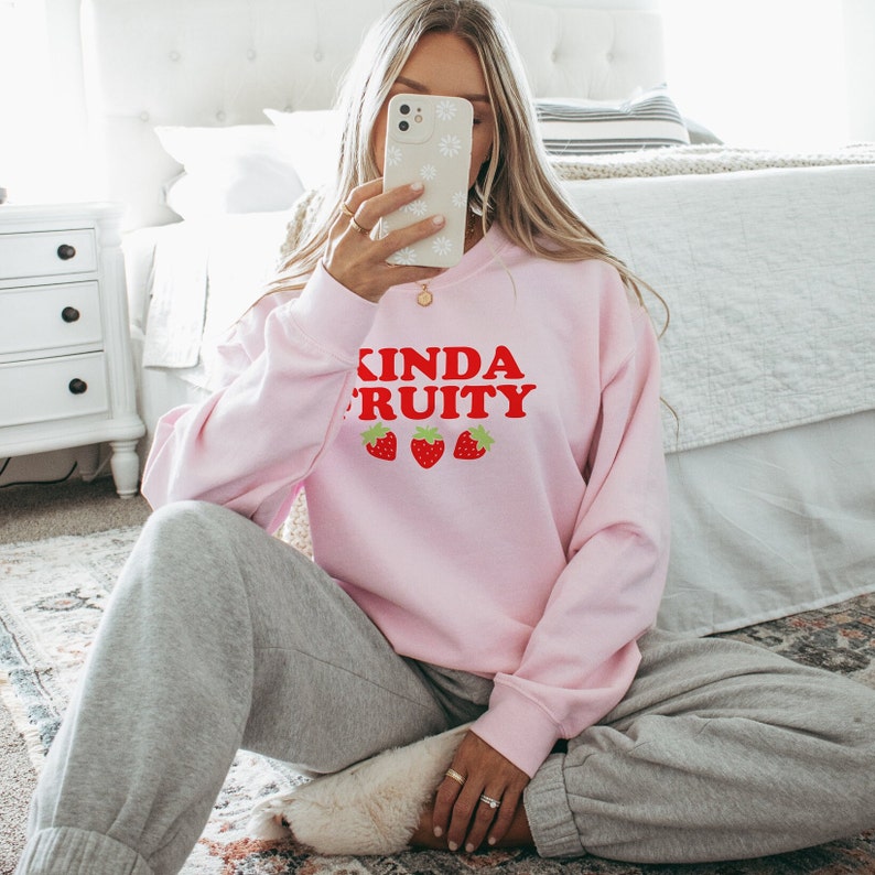 Puede incluir: Una sudadera rosa con el texto "Kinda Fruity" en letras rojas y tres fresas rojas impresas en la parte delantera.