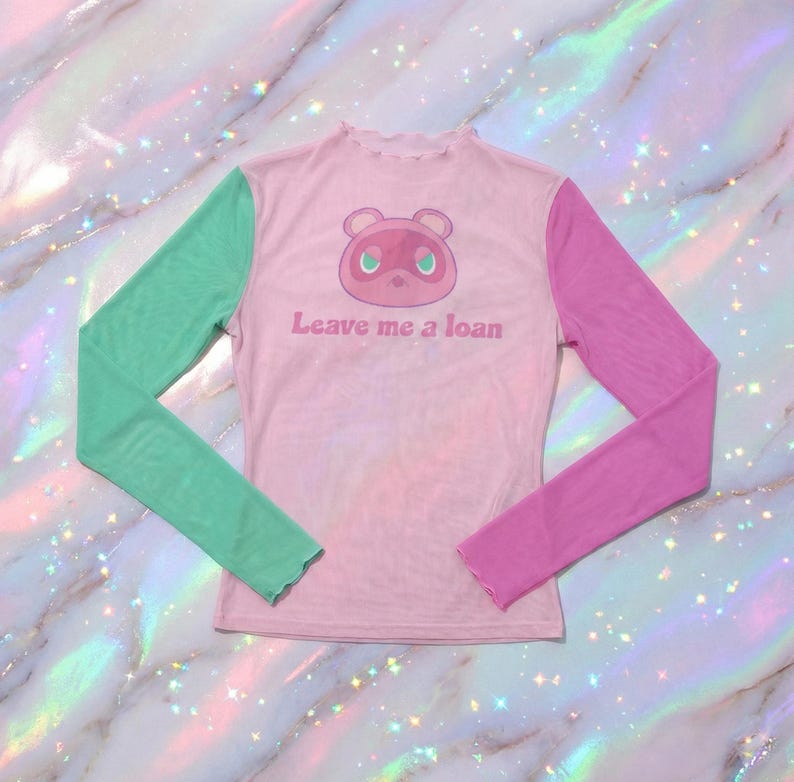 May include: A pink mesh long-sleeved top featuring a cartoon character and the text "Leave me a loan." The sleeves are green and purple. The top has a frilled neckline.