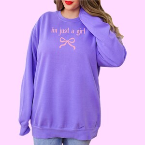Op de afbeelding: Paarse trui met de tekst "im just a girl" in roze met een roze strik eronder.