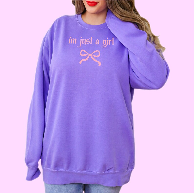 Op de afbeelding: Paarse trui met de tekst "im just a girl" in roze met een roze strik eronder.