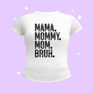 May include: A white short-sleeved t-shirt featuring black distressed-style text centered on the chest that reads MAMA. MOMMY. MOM. BRUH. The garment has a crew neckline and a fitted silhouette, presented against a light purple background with white stars.