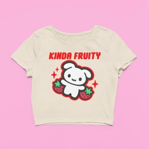 Op de afbeelding: Cr&egrave;mekleurige crop top met de tekst "KINDA FRUITY" in het rood. Het shirt heeft een cartoonbeer met aardbeien en rode steraccenten. Het ontwerp is gecentreerd op de voorkant van het shirt.