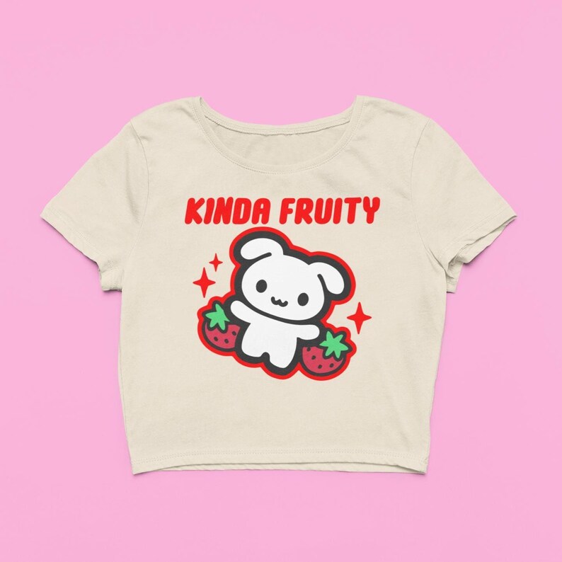 Op de afbeelding: Cr&egrave;mekleurige crop top met de tekst "KINDA FRUITY" in het rood. Het shirt heeft een cartoonbeer met aardbeien en rode steraccenten. Het ontwerp is gecentreerd op de voorkant van het shirt.