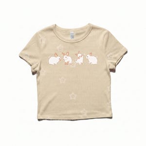 May include: A beige short-sleeved t-shirt featuring a graphic print of four small white rabbits in various poses across the chest. The fabric has a subtle, repeating star pattern throughout, and the rabbits are depicted with light brown ears.
