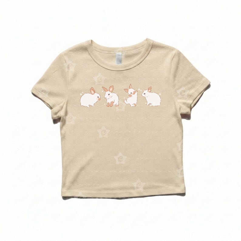May include: A beige short-sleeved t-shirt featuring a graphic print of four small white rabbits in various poses across the chest. The fabric has a subtle, repeating star pattern throughout, and the rabbits are depicted with light brown ears.