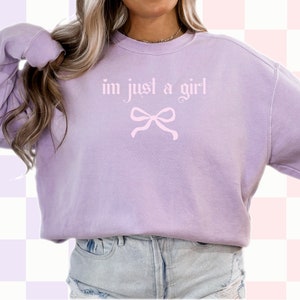 Op de afbeelding: Een lavendelkleurig sweatshirt met de tekst "im just a girl" in roze letters en een roze strik eronder.