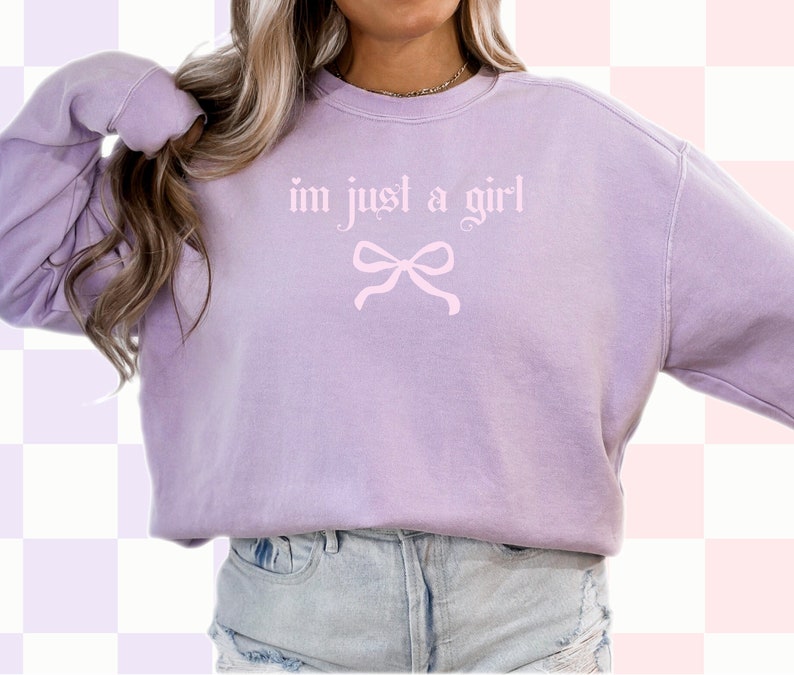 Op de afbeelding: Een lavendelkleurig sweatshirt met de tekst "im just a girl" in roze letters en een roze strik eronder.