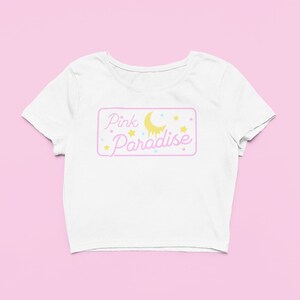 Pu&ograve; includere: Top corto bianco con grafica rettangolare rosa e gialla. La grafica mostra "Pink Paradise" in lettere rosa, una luna gialla e stelle. La maglietta ha maniche corte e scollo tondo.