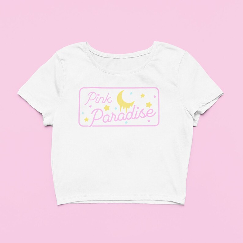 Pu&ograve; includere: Top corto bianco con grafica rettangolare rosa e gialla. La grafica mostra "Pink Paradise" in lettere rosa, una luna gialla e stelle. La maglietta ha maniche corte e scollo tondo.
