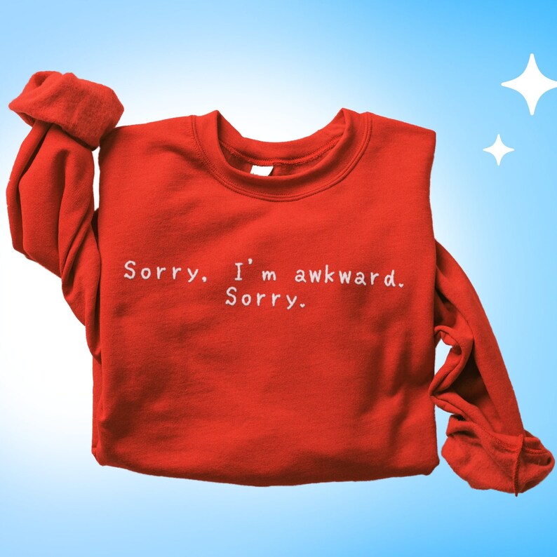 May include: A red sweatshirt with the text "Sorry. I'm awkward. Sorry." in white lettering. The sleeves are folded up. The background is a gradient of light blue to white with two white star shapes.
