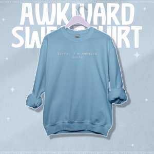 May include: A light blue sweatshirt with the text "Sorry. I'm awkward. Sorry." in a simple, sans-serif font. The word "AWKWARD SWEATSHIRT" is printed in large white letters above the sweatshirt.