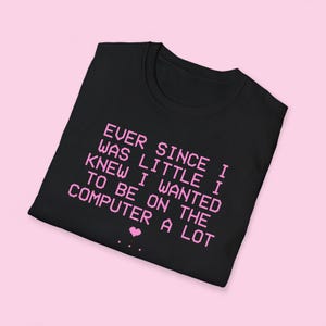 以下が含まれることがあります： 黒いTシャツには、EVER SINCE I WAS LITTLE I KNEW I WANTED TO BE ON THE COMPUTER A LOTと書かれた薄いピンク色のピクセル化されたテキストがあり、その下に小さなピンクのピクセル化されたハートと3つの点が中央に配置されています。