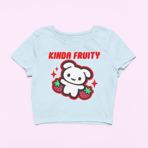 Op de afbeelding: Lichtblauw crop top met de rode tekst "Kinda Fruity". Het ontwerp bevat een witte cartoonbeer, twee aardbeien en rode steraccenten. Deze top is een casual item. Het shirt is een casual, alledaags kledingstuk.