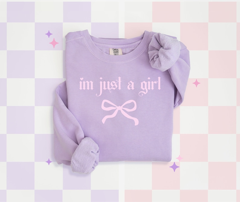 Op de afbeelding: Een lavendelkleurig sweatshirt met de tekst "im just a girl" in roze gotische letters en een roze strik. Het sweatshirt ligt op een geruite achtergrond.