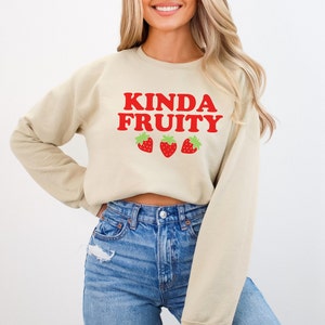 Puede incluir: Una sudadera corta de color beige claro con el texto "Kinda Fruity" en letras rojas y tres fresas rojas con hojas verdes.
