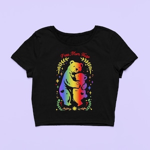 May include: Black crop top featuring a rainbow-colored bear hugging a smaller bear, with the text "Free Mom Hugs" in red script. The design is framed by a floral and star pattern. A cute and colorful graphic tee.
