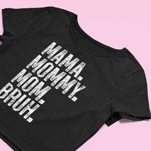 May include: A black short-sleeved t-shirt featuring white distressed-style text printed vertically on the front. The text reads: MAMA. MOMMY. MOM. BRUH. The garment has a round neckline and short sleeves, presented in a clean, straightforward manner.