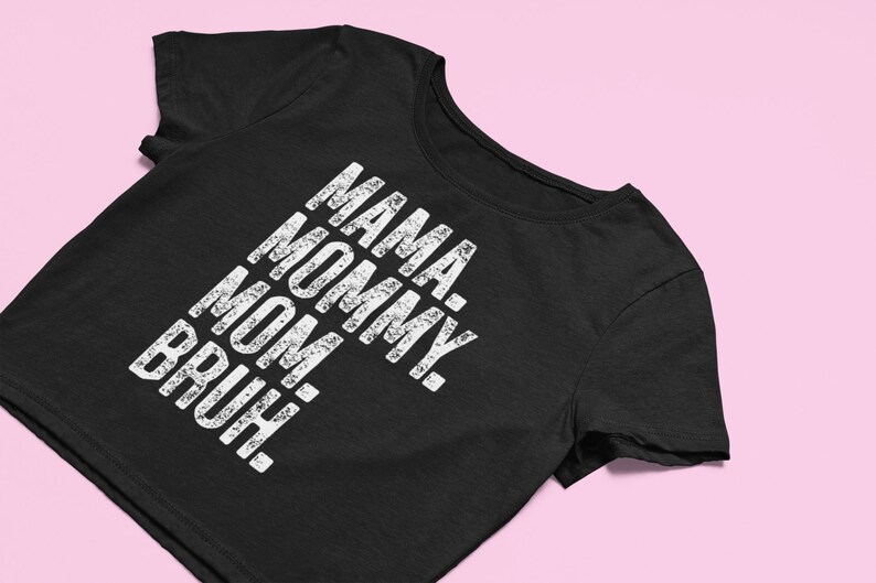 May include: A black short-sleeved t-shirt featuring white distressed-style text printed vertically on the front. The text reads: MAMA. MOMMY. MOM. BRUH. The garment has a round neckline and short sleeves, presented in a clean, straightforward manner.