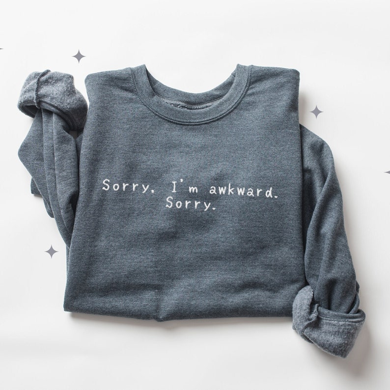 May include: A dark gray sweatshirt with the text "Sorry. I'm awkward. Sorry." in white. The long sleeves are rolled up. The sweatshirt is displayed on a white surface with star-shaped decorations.