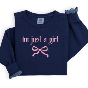 Op de afbeelding: Navyblauwe trui met een roze strik en de tekst "i'm just a girl" in gotische letters.