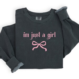 Op de afbeelding: Een donkergrijs sweatshirt met de tekst "im just a girl" in roze gotische letters en een roze strik eronder.