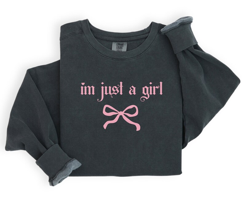 Op de afbeelding: Een donkergrijs sweatshirt met de tekst "im just a girl" in roze gotische letters en een roze strik eronder.