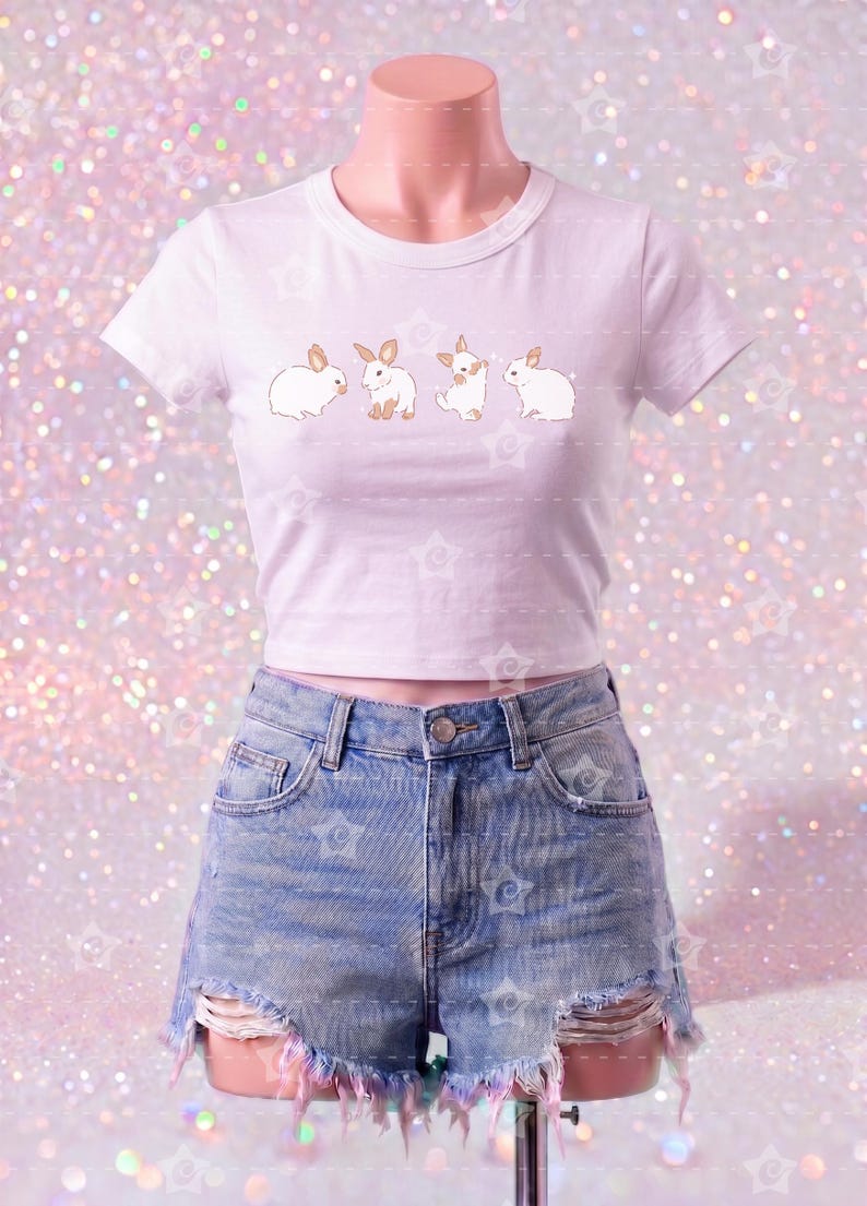 May include: A white short-sleeved crop top featuring a horizontal row of four small brown and white rabbit illustrations across the chest, paired with light blue denim shorts that have a distressed, frayed hemline and visible pocket lining.