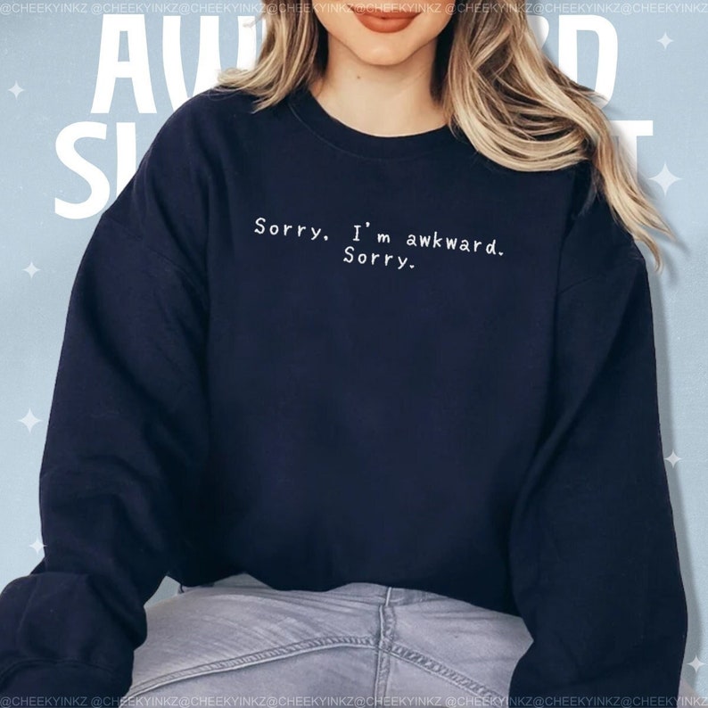 May include: Navy blue sweatshirt with the text "Sorry. I'm awkward. Sorry." in white. The sweatshirt is a crew neck style and has long sleeves. The text is centered on the chest.