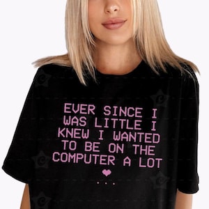 May include: A black short-sleeved t-shirt featuring the light pink pixelated text: EVER SINCE I WAS LITTLE I KNEW I WANTED TO BE ON THE COMPUTER A LOT, followed by a small pink pixelated heart and three dots centered below the text.