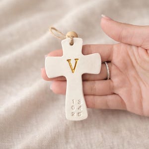 Puede incluir: Un colgante de cruz de cerámica blanca con la letra dorada "V" y los números "13 04 25" en relieve. Tiene un lazo de cuerda natural y una cuenta de madera para colgar. La cruz se sostiene sobre un fondo de tela neutra.