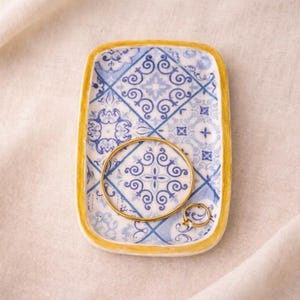 Puede incluir: Una bandeja rectangular de cerámica con un patrón de azulejos azules y blancos y un borde dorado. Una pulsera de brazalete dorada y un anillo dorado descansan dentro de la bandeja. La bandeja está sobre una superficie color crema.