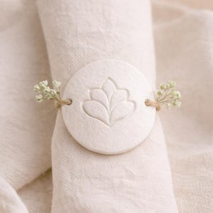 Puede incluir: Un servilletero blanco y circular con un diseño floral, sujeto con hilo y pequeñas flores blancas. El servilletero está hecho de un material mate y está envuelto alrededor de una servilleta de lino doblada.