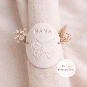 Puede incluir: Un servilletero de cerámica blanca con la palabra "MAMA" y un diseño de mariposa. El anillo está adornado con pequeñas flores secas y el texto "Can be personalised". El servilletero está alrededor de una servilleta de lino blanca.